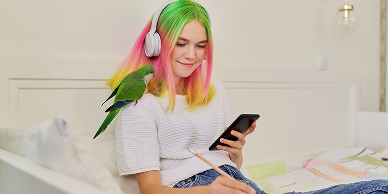 teenage girl sitting on a bed doing homework with headphones and a mobile phone with a pet parrot on her shoulder